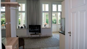 A modern living room with a flat-screen TV, a dining area with a wooden table, and large windows with curtains. - Haus Wanda Appartement 4a - Haus Wanda (Heringsdorf)