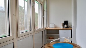 A small kitchenette with a toaster, coffee maker, and a refrigerator. There is a chair with a blue mat and a window with a view of trees. - House Wanda Apartment 05 - House Wanda (Heringsdorf)