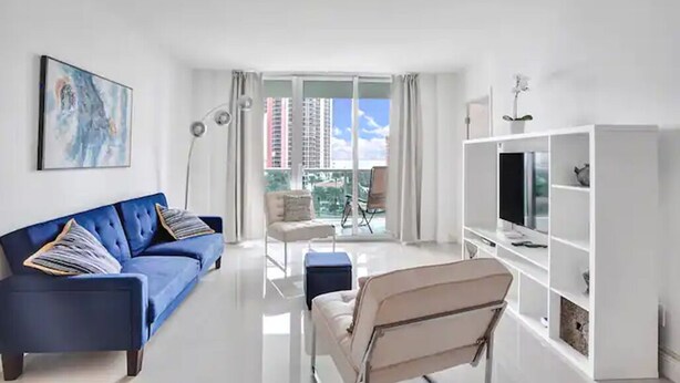 Un salon moderne avec un canapé bleu, un fauteuil blanc, un téléviseur posé sur un meuble blanc et une grande fenêtre donnant sur des immeubles.