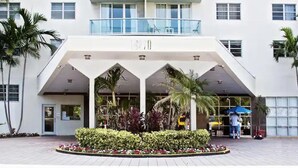 L’entrée d’un hôtel, avec un espace abrité, des palmiers et un parterre de fleurs.