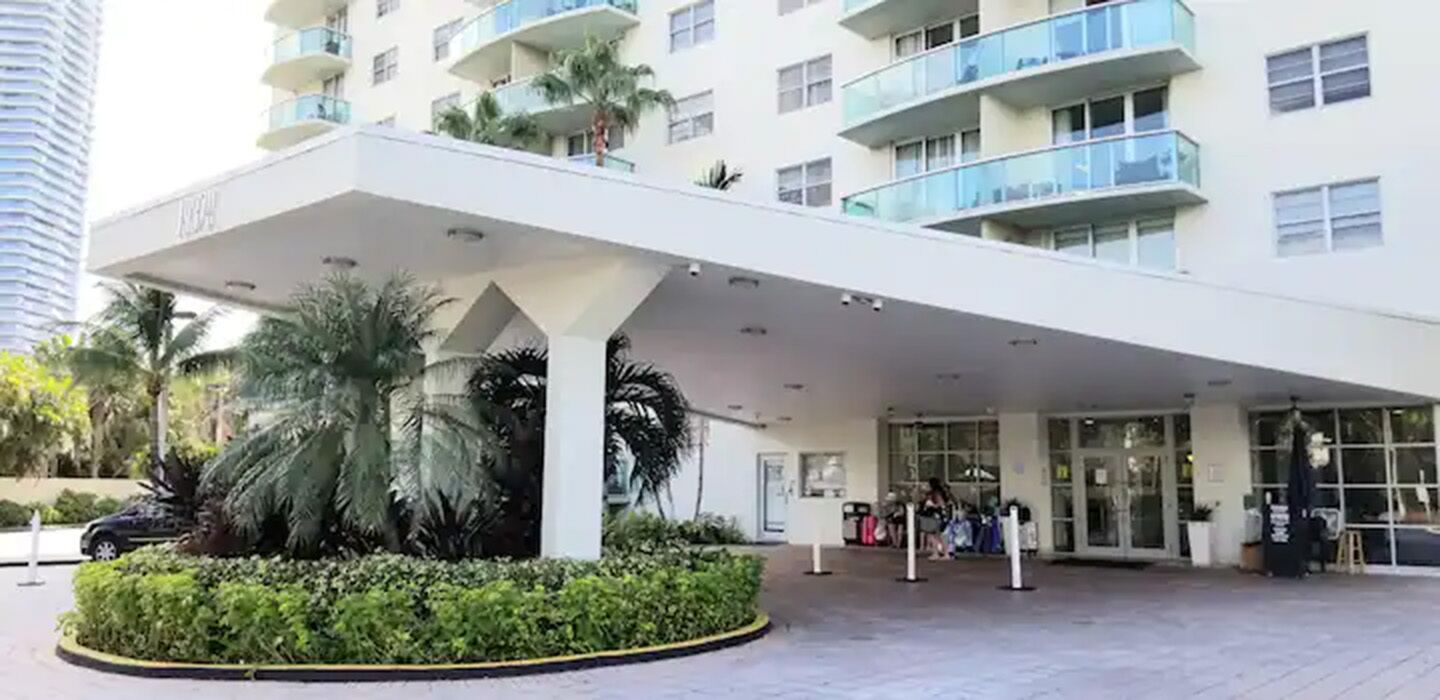 Une entrée d’hôtel moderne, dotée d’une allée couverte, de palmiers et d’une façade en verre.