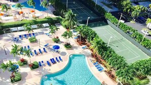 Vue aérienne d’un complexe hôtelier doté d’une piscine, de chaises longues, d’un court de tennis et de palmiers.