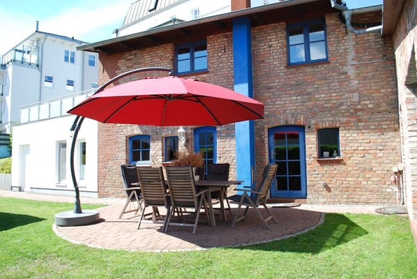 Un patio con una mesa y sillas, un paraguas rojo y un edificio de ladrillo con puertas azules.