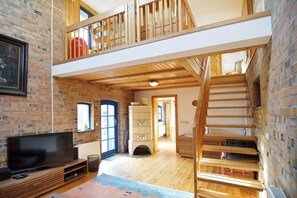Un espacio tipo loft con vigas de madera a la vista, pared de ladrillo, televisor y escalera que sube a un nivel superior.