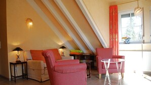 Habitación estilo loft con techo inclinado, un sofá, dos sillones, una mesita redonda y una ventana con cortinas.