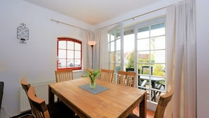 Ein Essbereich mit einem Holztisch, sechs Stühlen, einer Blumenvase, einem Fenster mit Vorhängen und einem Heizkörper.