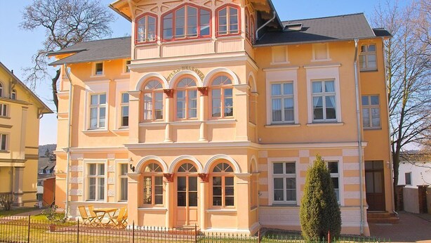 Ein mehrstöckiges Gebäude mit einer symmetrischen Fassade, großen Fenstern und einem markanten Balkon.