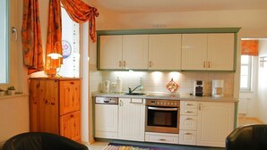 Eine kompakte Küche mit weißen Schränken, einem Geschirrspüler, einem Backofen und einer Kaffeemaschine. Links befindet sich ein Holzschrank und im Vordergrund ein schwarzer Stuhl.