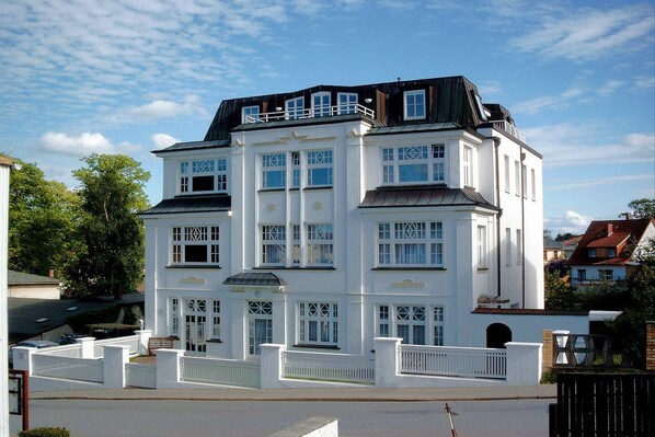 Ein mehrstöckiges weißes Gebäude mit einem Balkon im zweiten Stock, großen Fenstern und einem verschlossenen Eingang.