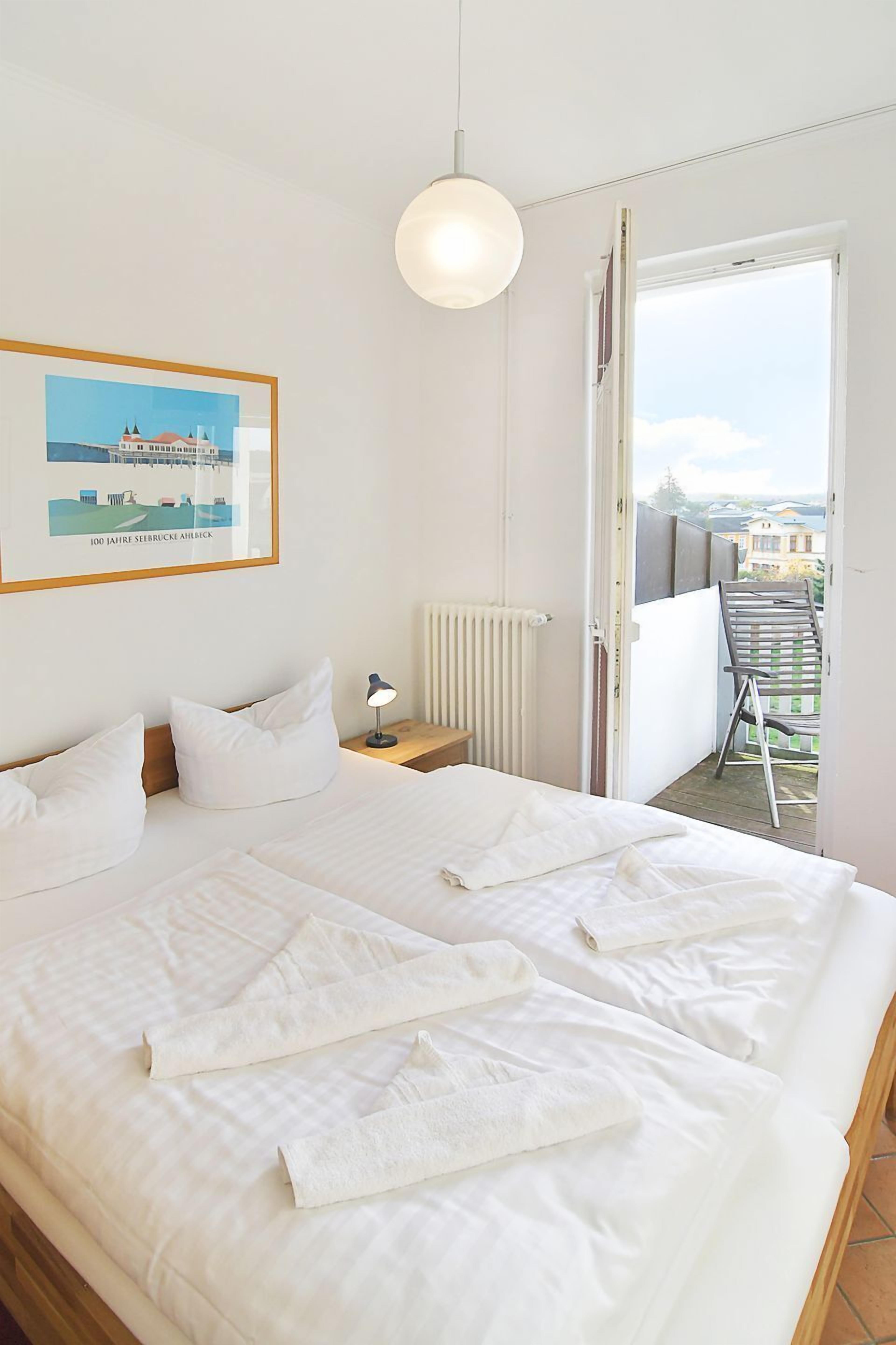 A hotel room with a large bed, a bedside table with a lamp, a framed picture on the wall, and a balcony with a chair.