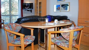 Een eetgedeelte met een ronde tafel, twee stoelen, een zwarte leren bank en een aan de muur bevestigde televisie.