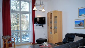 Una sala de estar con televisor, un mueble de madera, un sofá de cuero negro y una ventana con cortinas rojas.