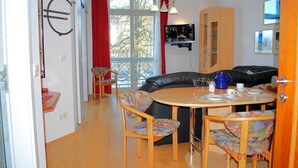 Een gezellige eethoek met een ronde tafel, stoelen en een bank. Een televisie is aan de muur bevestigd en er hangen gordijnen voor de ramen.