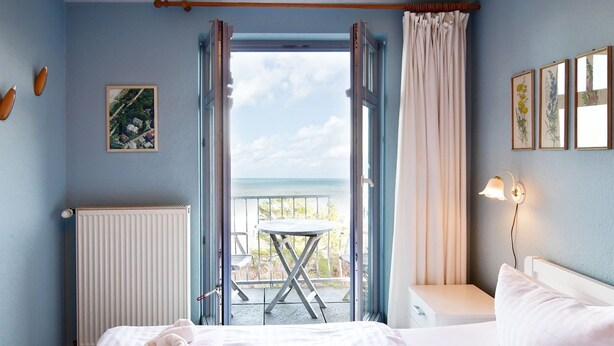 A bedroom with a bed, a bedside table, a window with curtains, a radiator, and a balcony with a table and chairs.