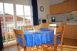 Un coin repas avec une table recouverte d’une nappe bleue, des chaises en bois, un panier en osier et une fenêtre donnant sur les bâtiments à l’extérieur.