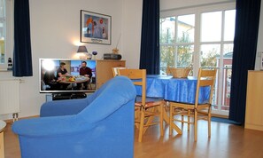 Ein Wohnzimmer mit Fernseher, blauem Sofa, Esstisch mit Stühlen und einem Fenster mit Vorhängen.