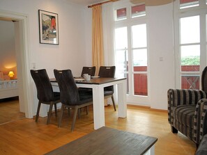 A dining area with a table and chairs, a window with curtains, and a view of greenery outside. - Villa Vineta Appartement 15 - Villa Vineta (Heringsdorf)