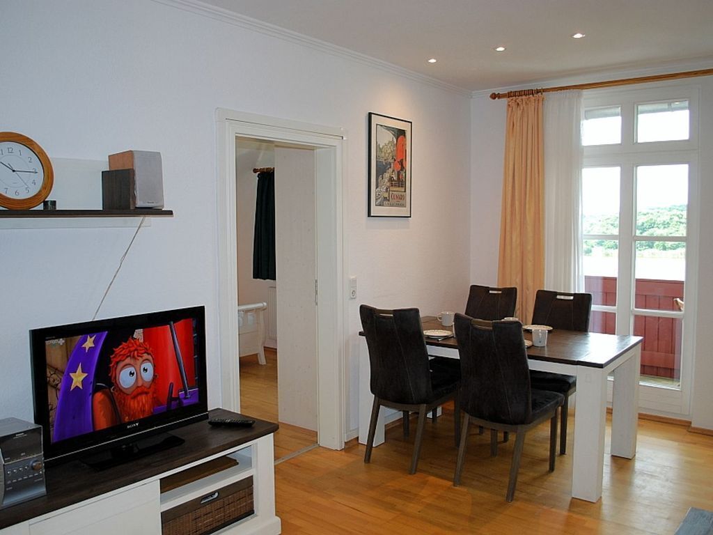 A living room with a TV, a dining table with chairs, a clock, and a window with curtains.