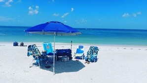Une plage avec un parasol bleu, deux chaises de plage, une glacière et une bouteille d’eau.