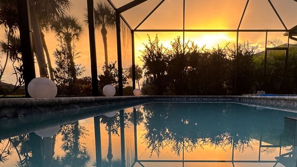 Une piscine entourée de parois en verre qui reflètent le coucher de soleil et les palmiers environnants.