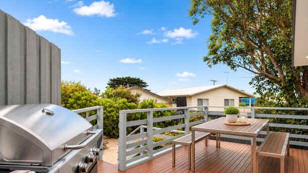 A deck with a barbecue grill, a table with a candle, and a view of a house and greenery.