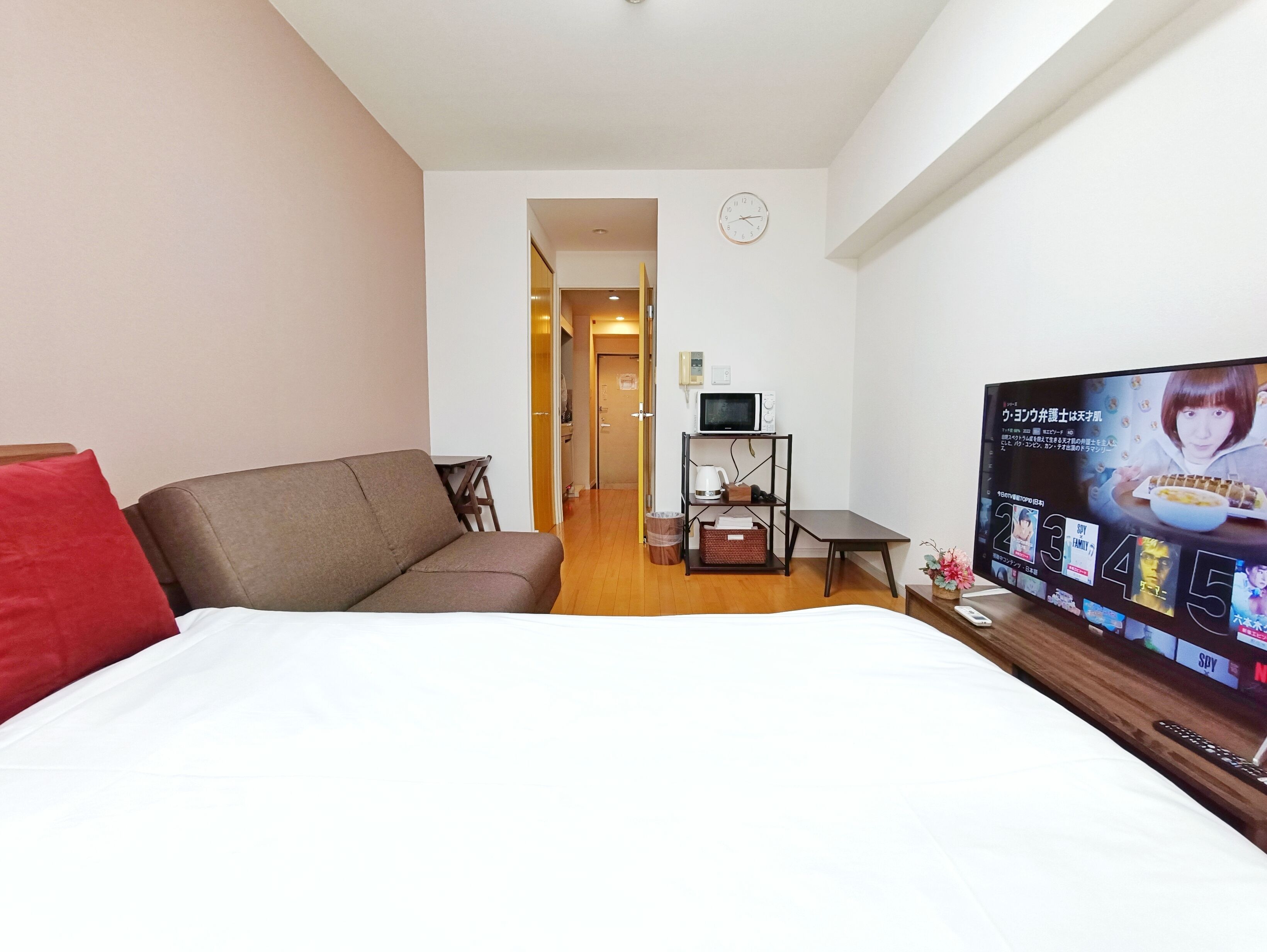 Quarto de hotel com cama, sofá marrom, televisão e uma pequena mesa com chaleira e cesta.