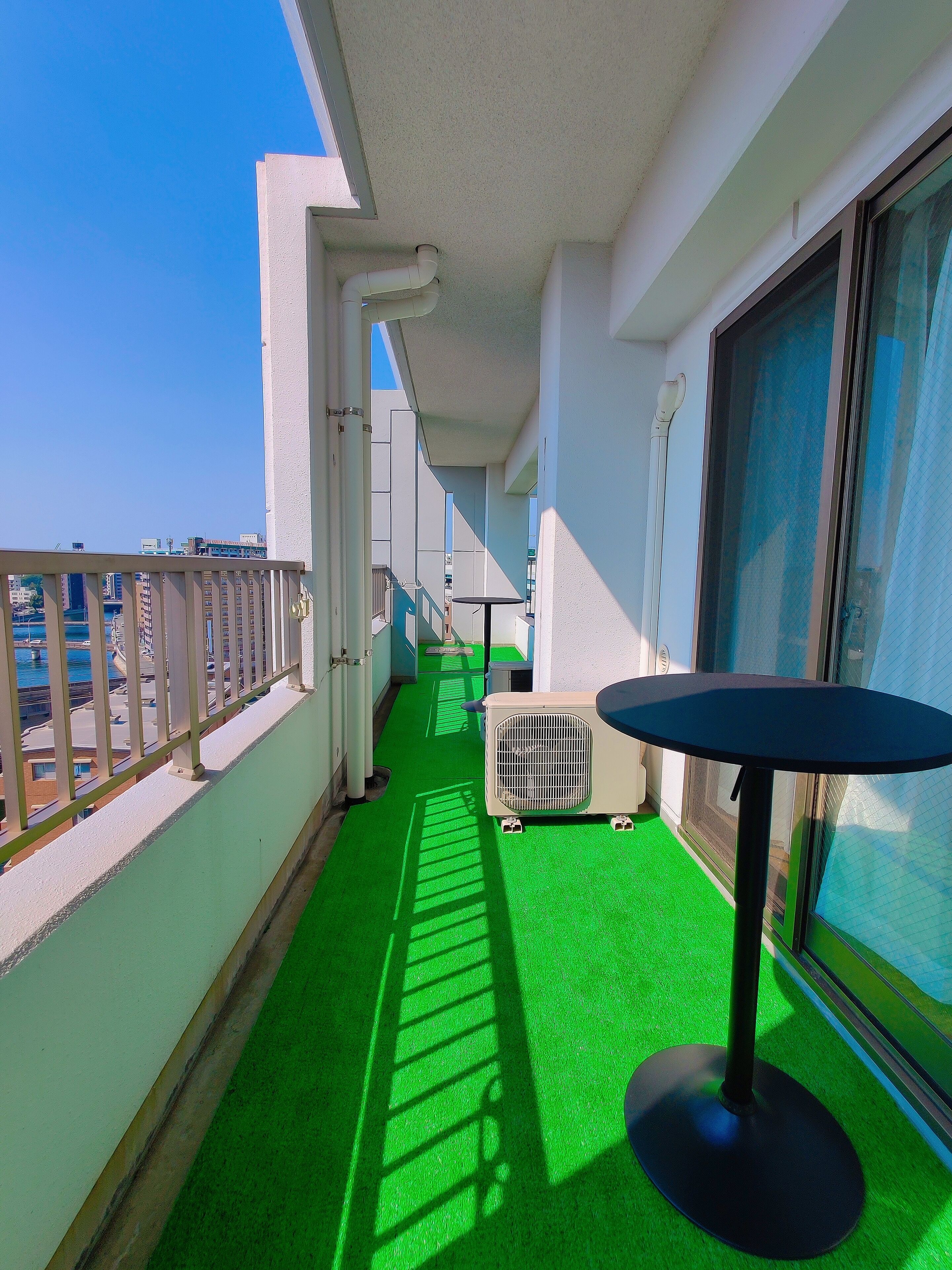 Un balcon doté d’un sol recouvert d’un gazon synthétique vert, d’une petite table et d’une fenêtre donnant sur des immeubles.