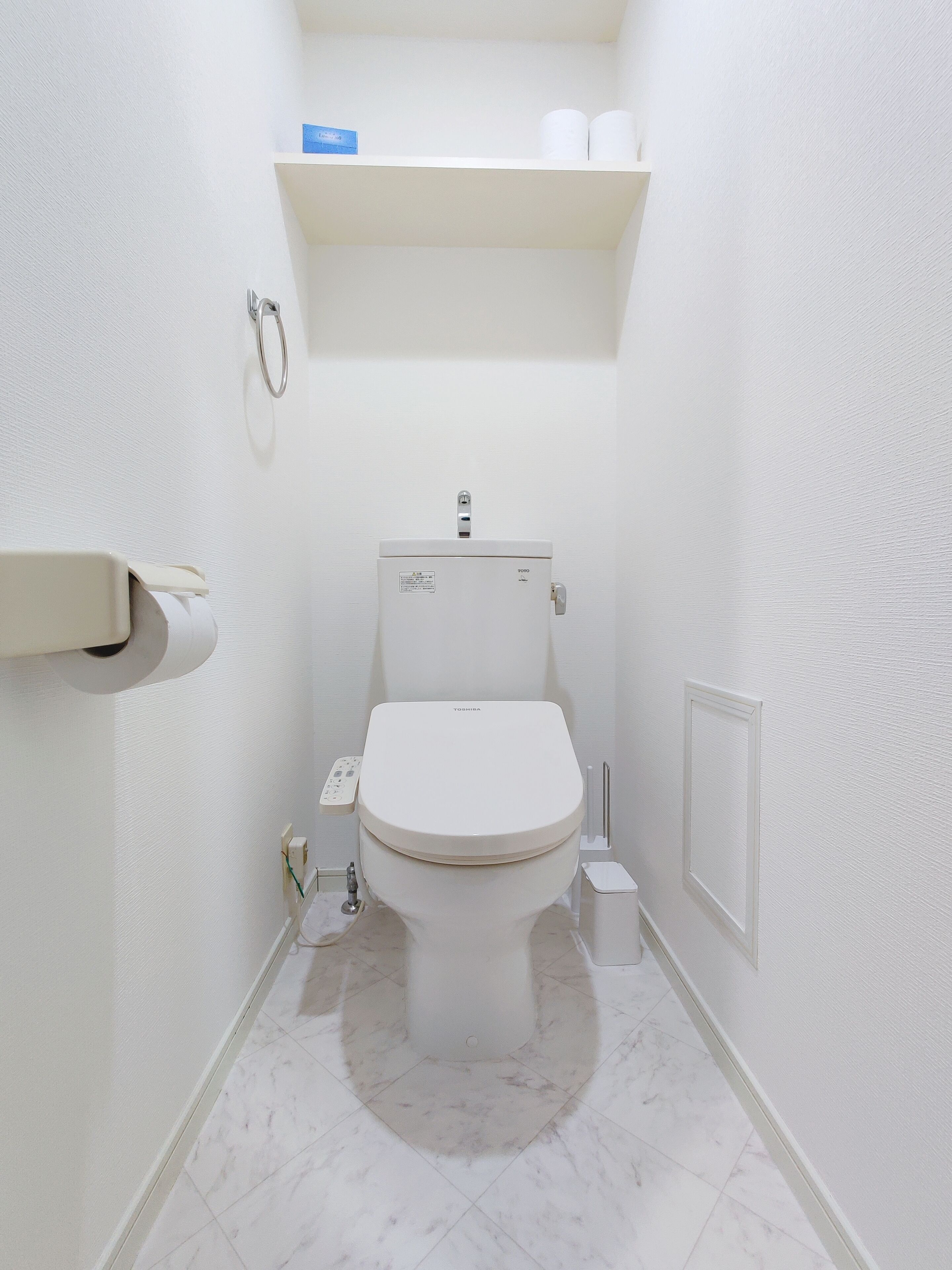 Un baño pequeño y blanco con inodoro, dispensador de toallas de papel y repisa con papel higiénico.