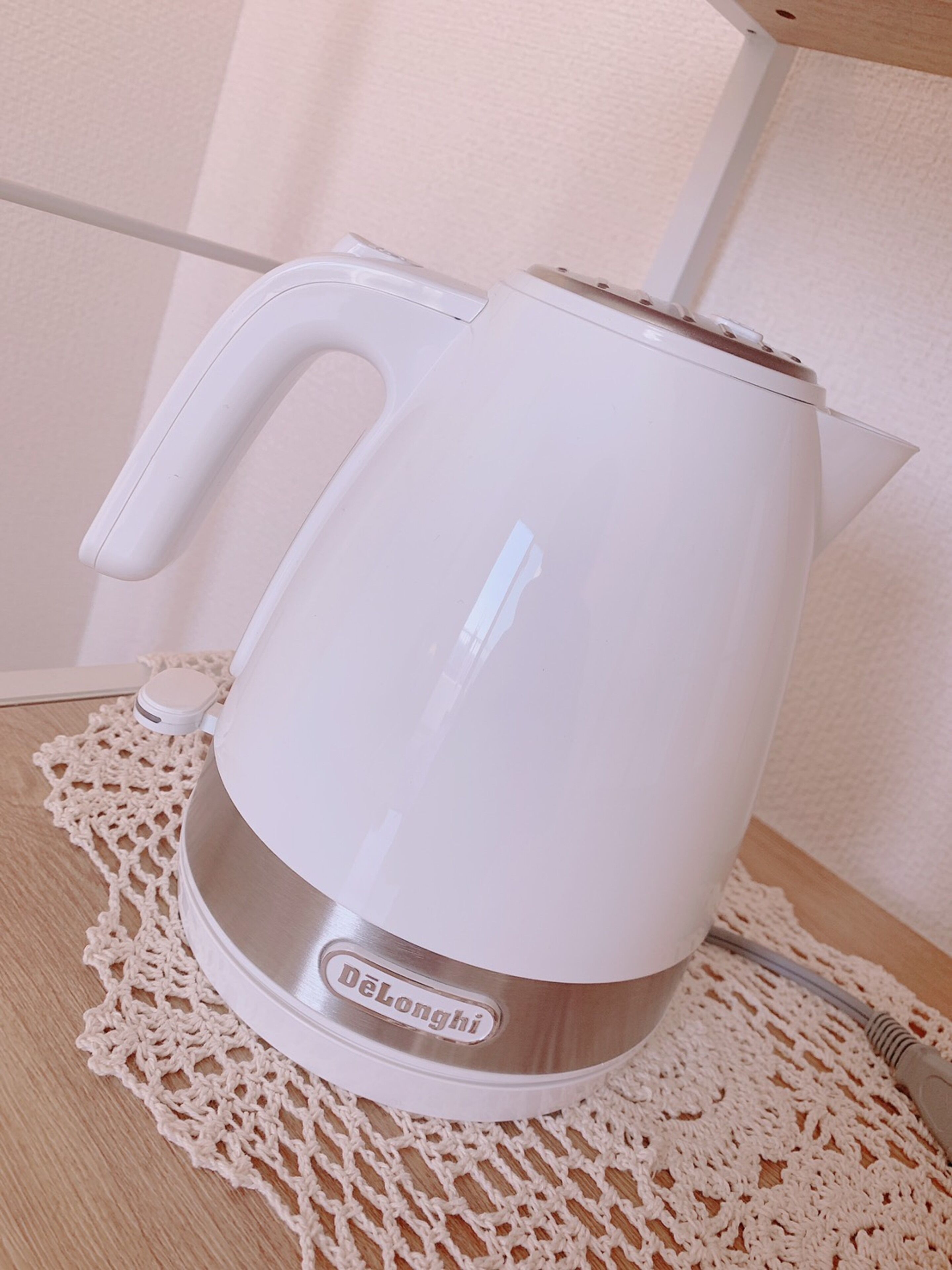 A white electric kettle with a handle and a base, placed on a wooden surface with a crocheted doily.