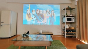 A living room with a flat-screen TV mounted on the wall, a glass-top coffee table, a microwave, and a bookshelf.