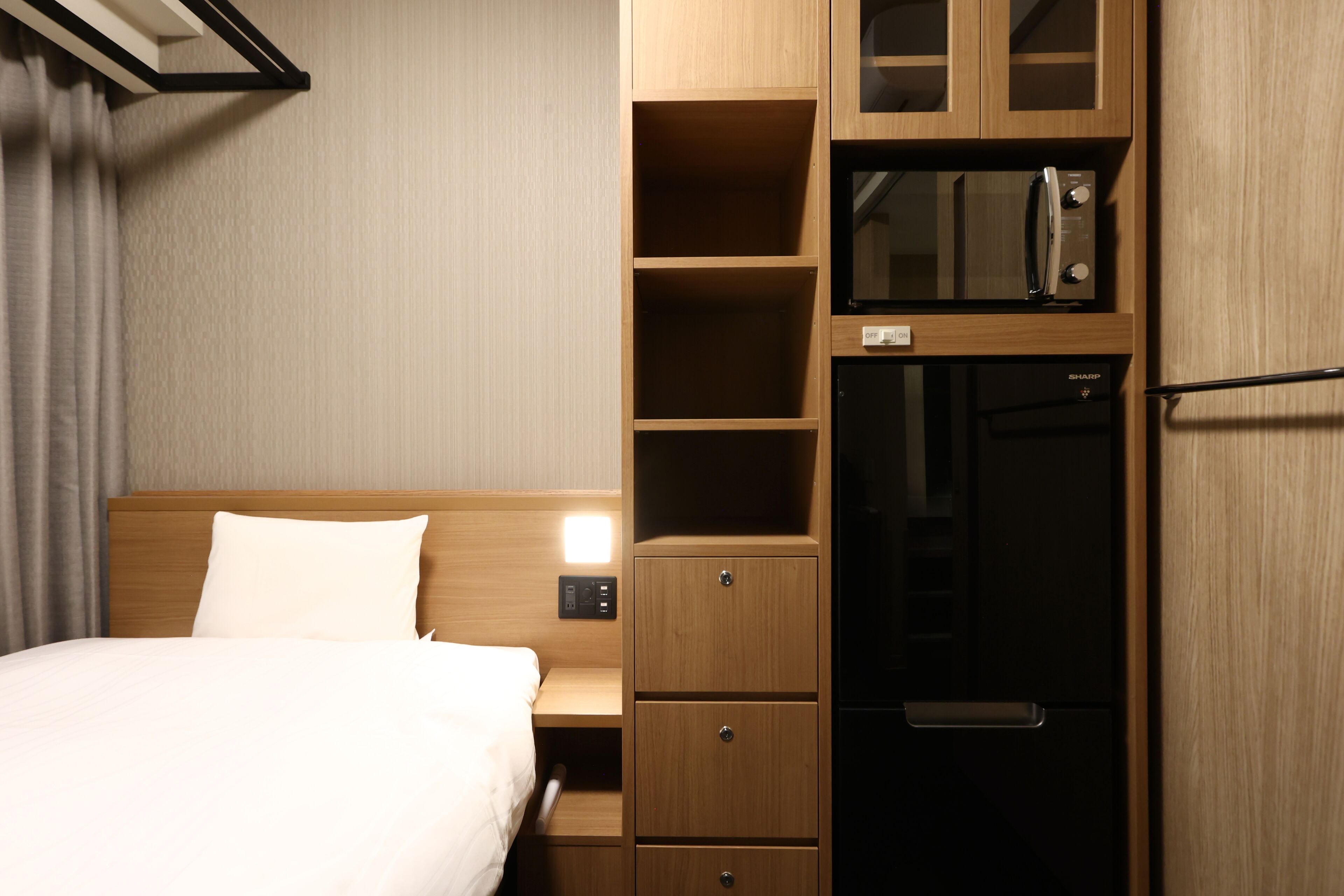 A hotel room with a bed, wooden wardrobe, built-in microwave, and a refrigerator.