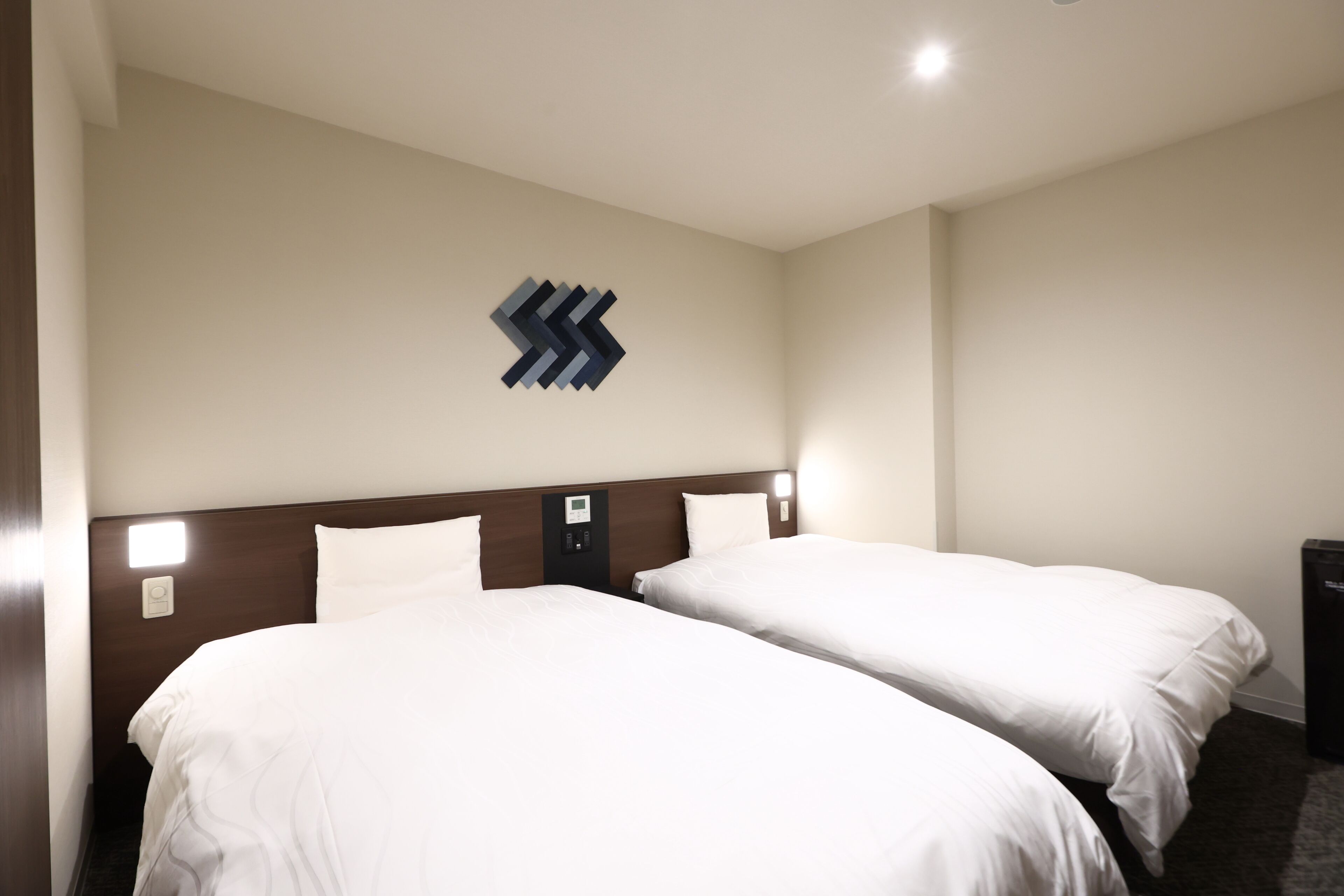 A hotel room with two beds, a wooden headboard, a wall-mounted light, and a small trash bin.