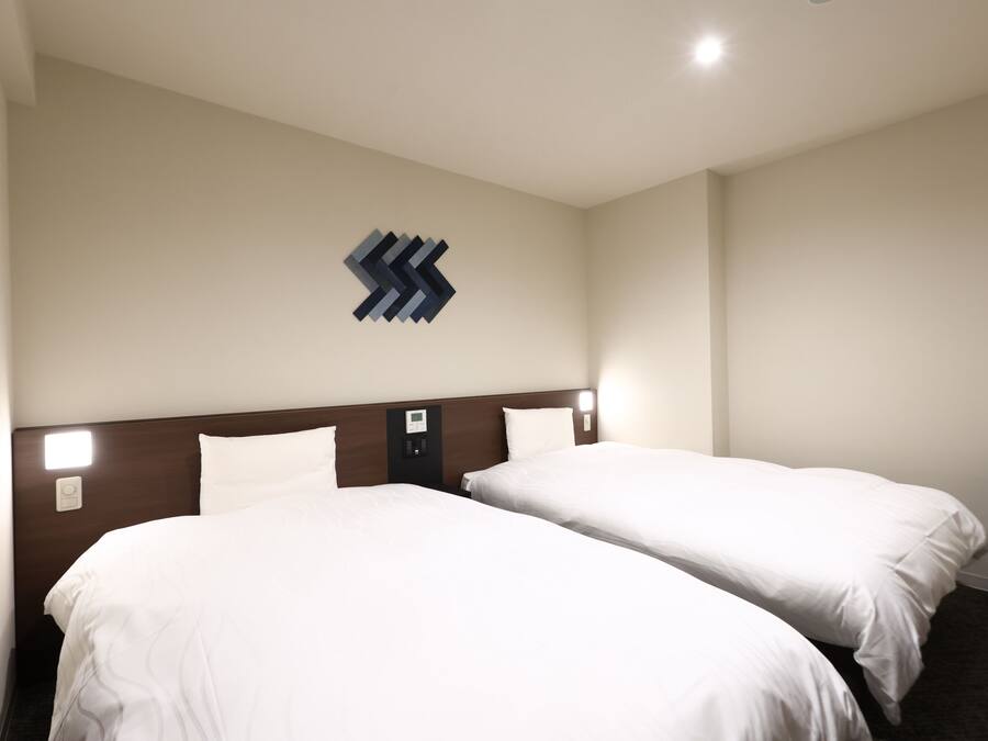 Habitación de hotel con dos camas, cabecera de madera, luz de pared y un pequeño cesto de basura.