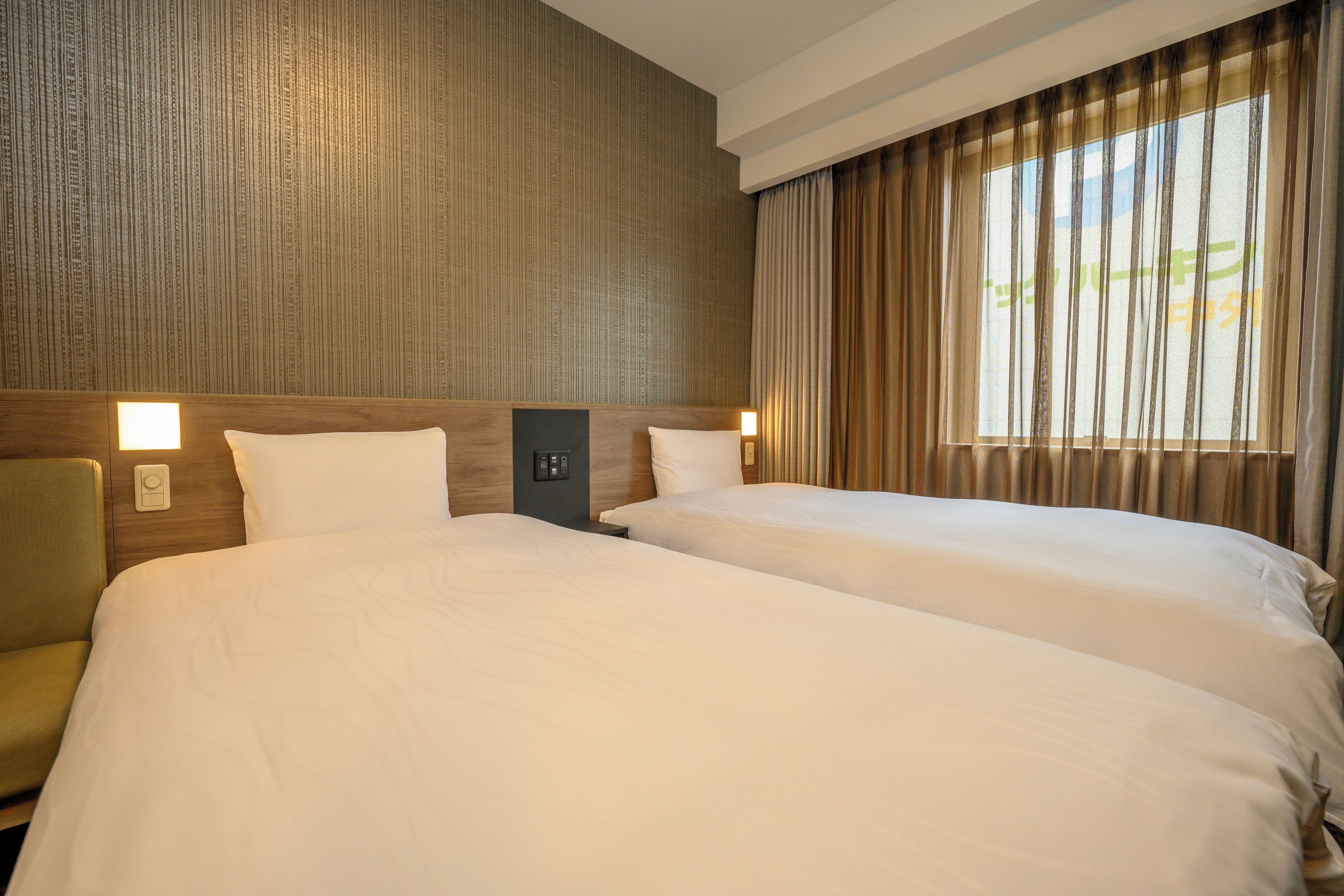 Habitación de hotel con dos camas, cabecera de madera, mesita de noche y ventana con cortinas.