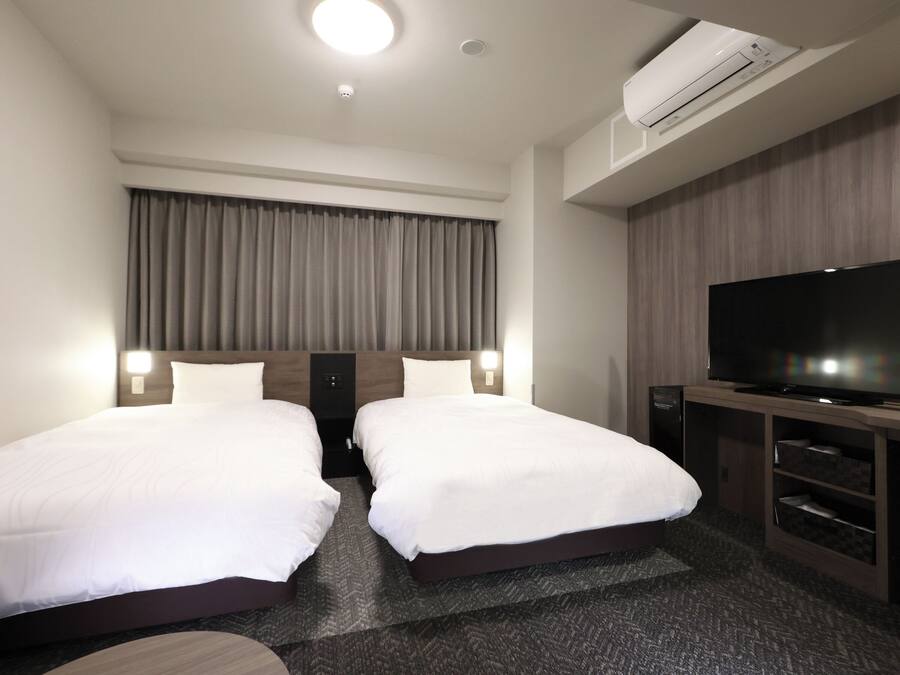 A hotel room with two single beds, a flat-screen TV, a built-in air conditioning unit, and a large window with curtains.