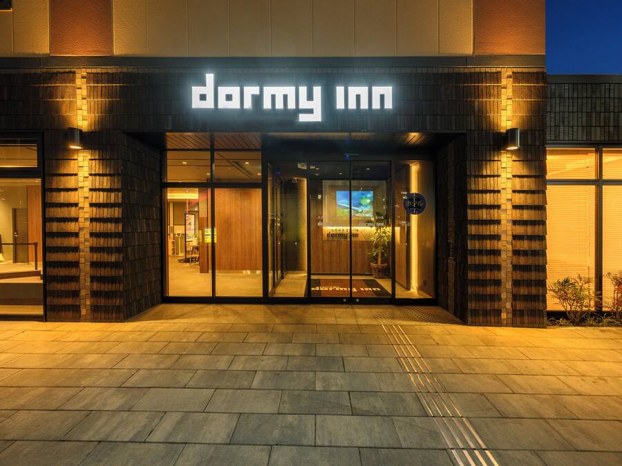 The exterior of a hotel named 'Dormy Inn' with a modern design, featuring large glass doors and a welcoming entrance.