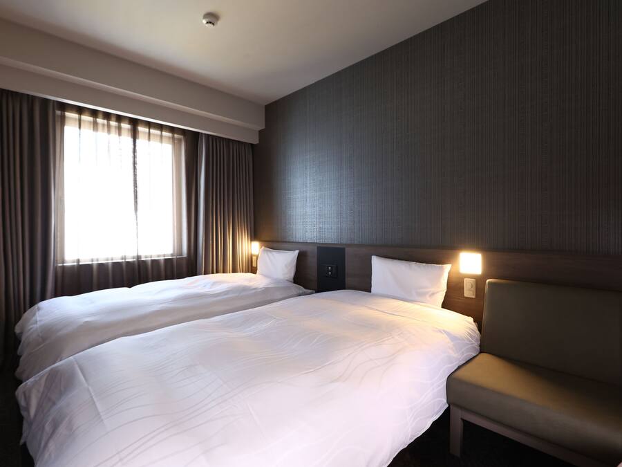 Une chambre d’hôtel avec deux lits, un canapé, une grande fenêtre avec des rideaux et des lampes fixées au mur.