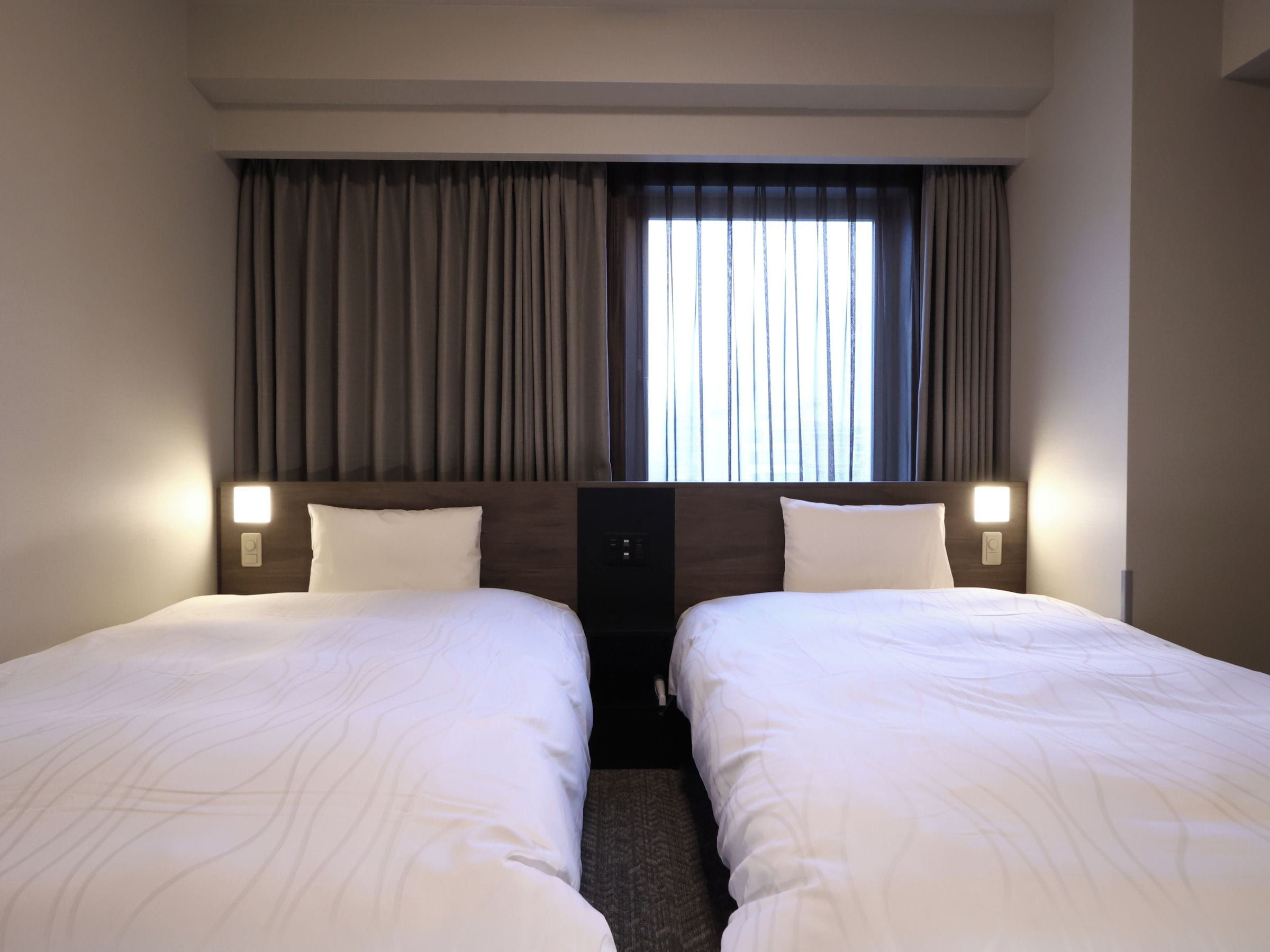 A hotel room with two single beds, a large window with curtains, and bedside lamps.