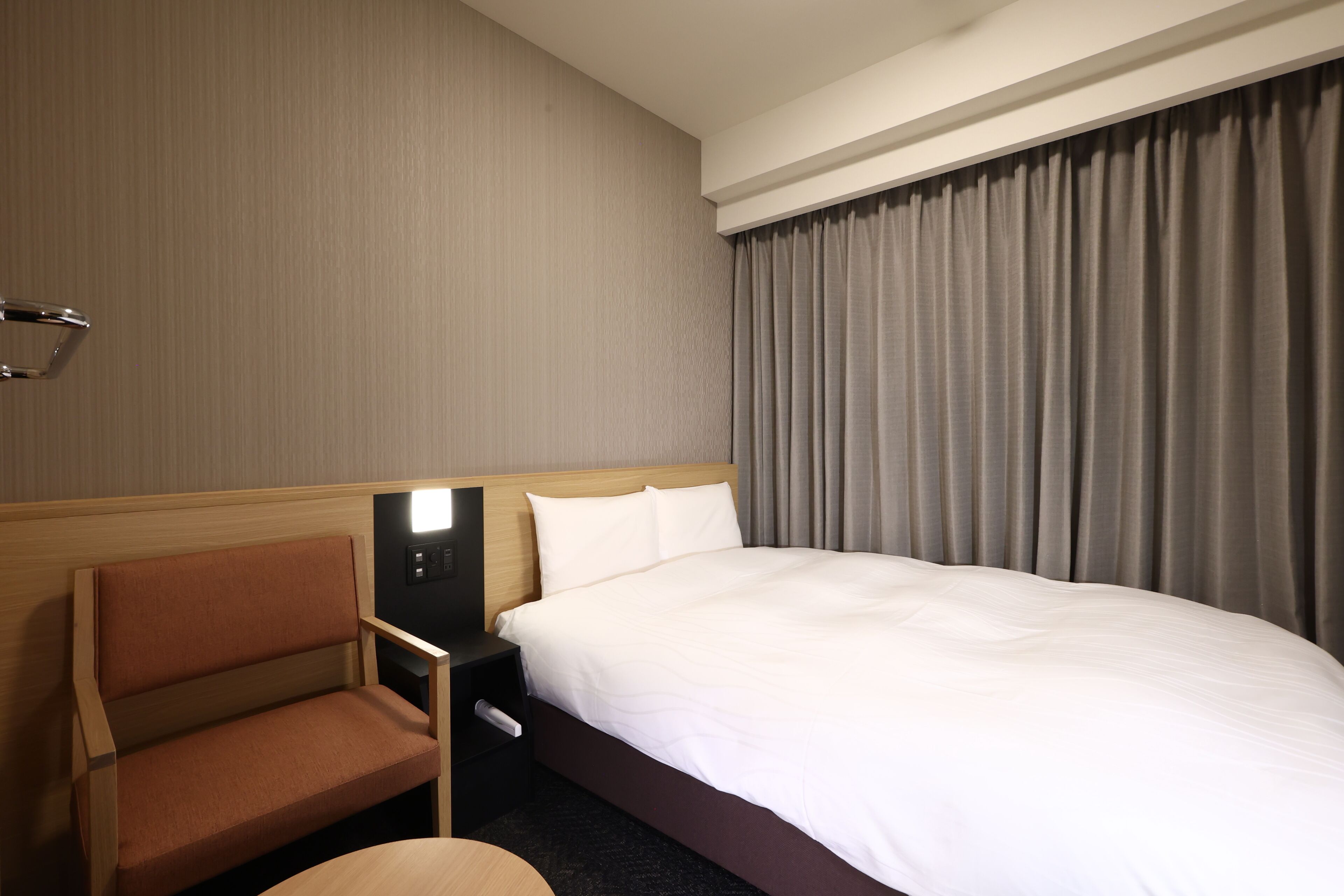 Quarto de hotel com uma cama, mesa de cabeceira, cadeira e janela com cortinas.