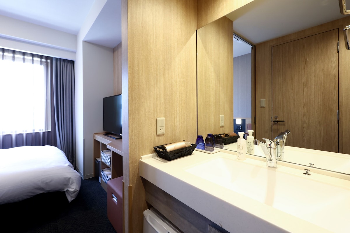 A hotel room with a large bed, a TV on a stand, a wooden wardrobe, a bathroom with a sink and mirror, and a window with curtains.