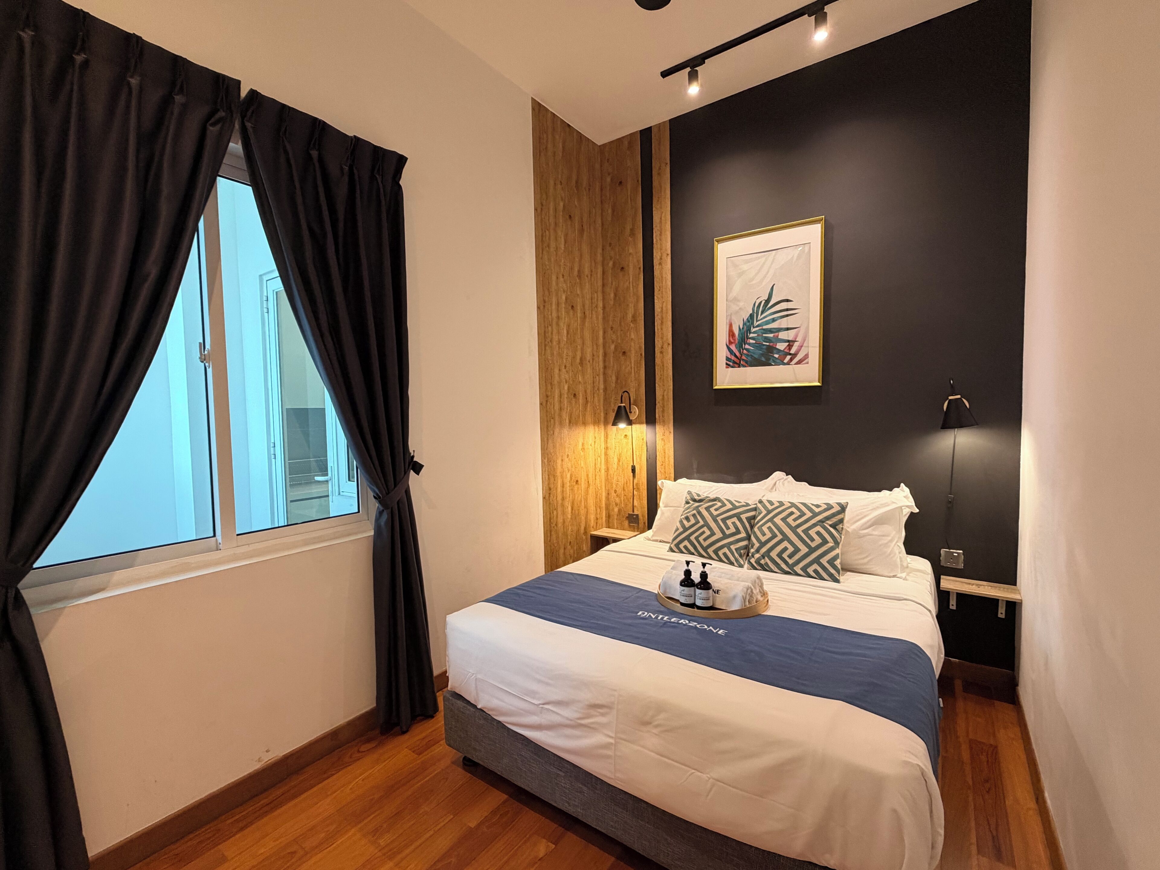 A hotel room with a bed, a window with curtains, a wall-mounted lamp, and a framed picture.
