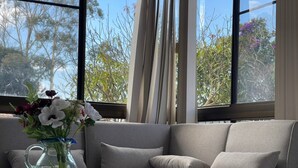 A modern living room with a grey sofa, a glass vase with flowers, and a view of trees through large windows. - Modern than 3 bedroom house  with office space in the center of Kallangur (Kallangur)