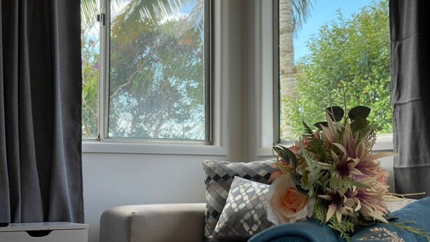 A bedroom with a bed, a window with curtains, a view of palm trees, and a floral arrangement on the bed. - Modern than 3 bedroom house  with office space in the center of Kallangur (Kallangur)