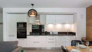 A modern kitchen with white cabinets, a black oven, and a wooden dining table set with wine glasses and plates. - Apartment Quintessence - B101 (CHATEL)