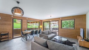 A modern living room with a sofa, dining table, and chairs, large windows, and a TV. - Apartment Quintessence - B101 (CHATEL)