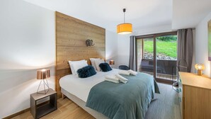 A modern bedroom with a wooden headboard, a large bed with a green bedspread, two decorative pillows, a bedside lamp, and a view of a garden through a sliding glass door. - Apartment Quintessence - B101 (CHATEL)