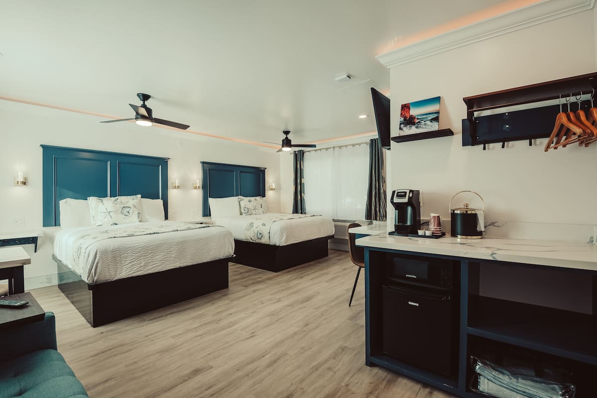 A modern hotel room with two beds, a kitchenette, a ceiling fan, and a mounted TV.