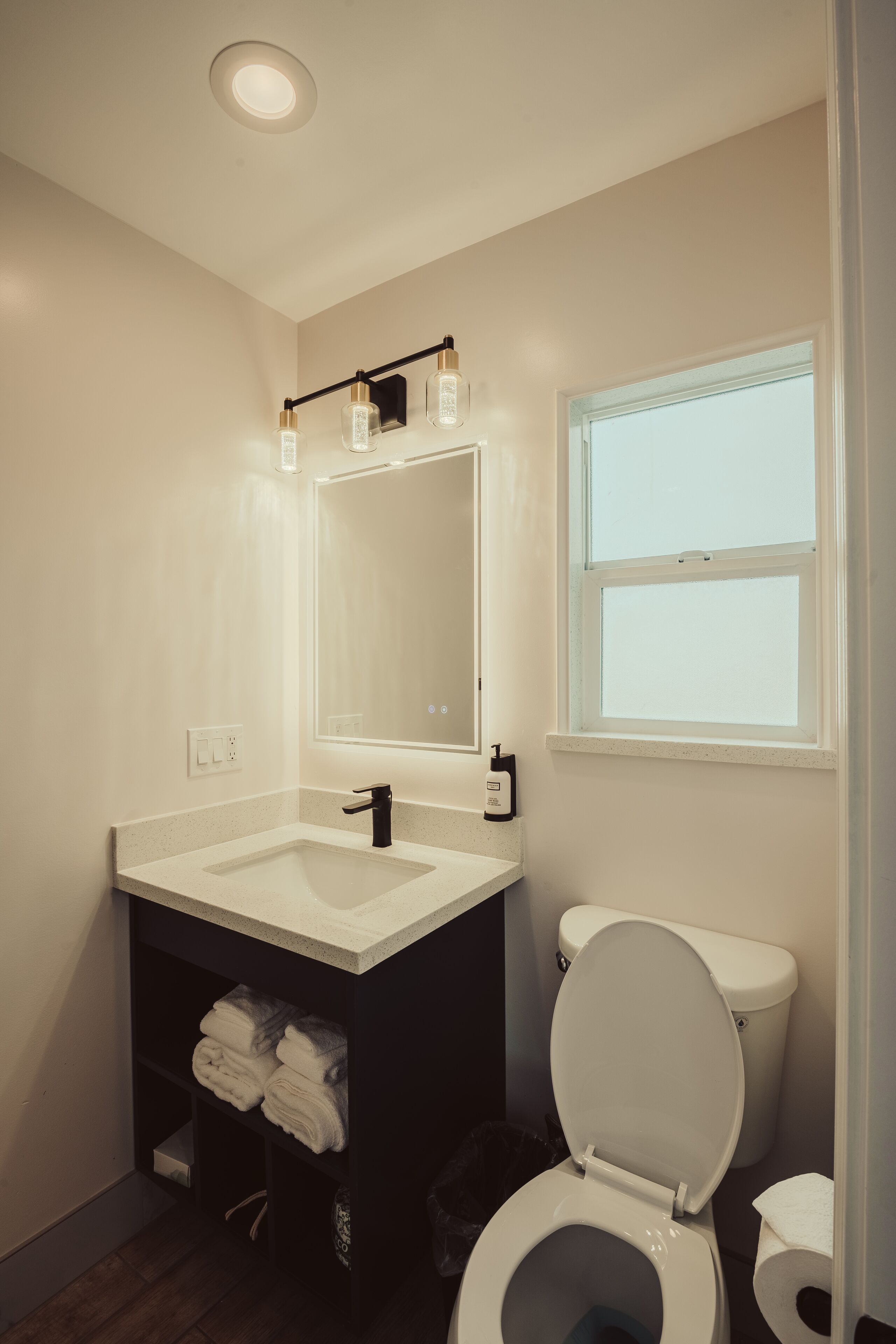 A modern bathroom with a white sink, black vanity, and a toilet. There is a window providing natural light and a mirror above the sink.