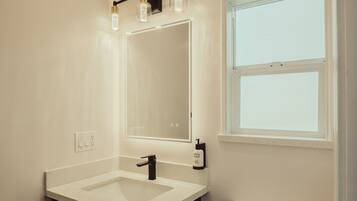 A modern bathroom with a white sink, black vanity, and a toilet. There is a window providing natural light and a mirror above the sink.
