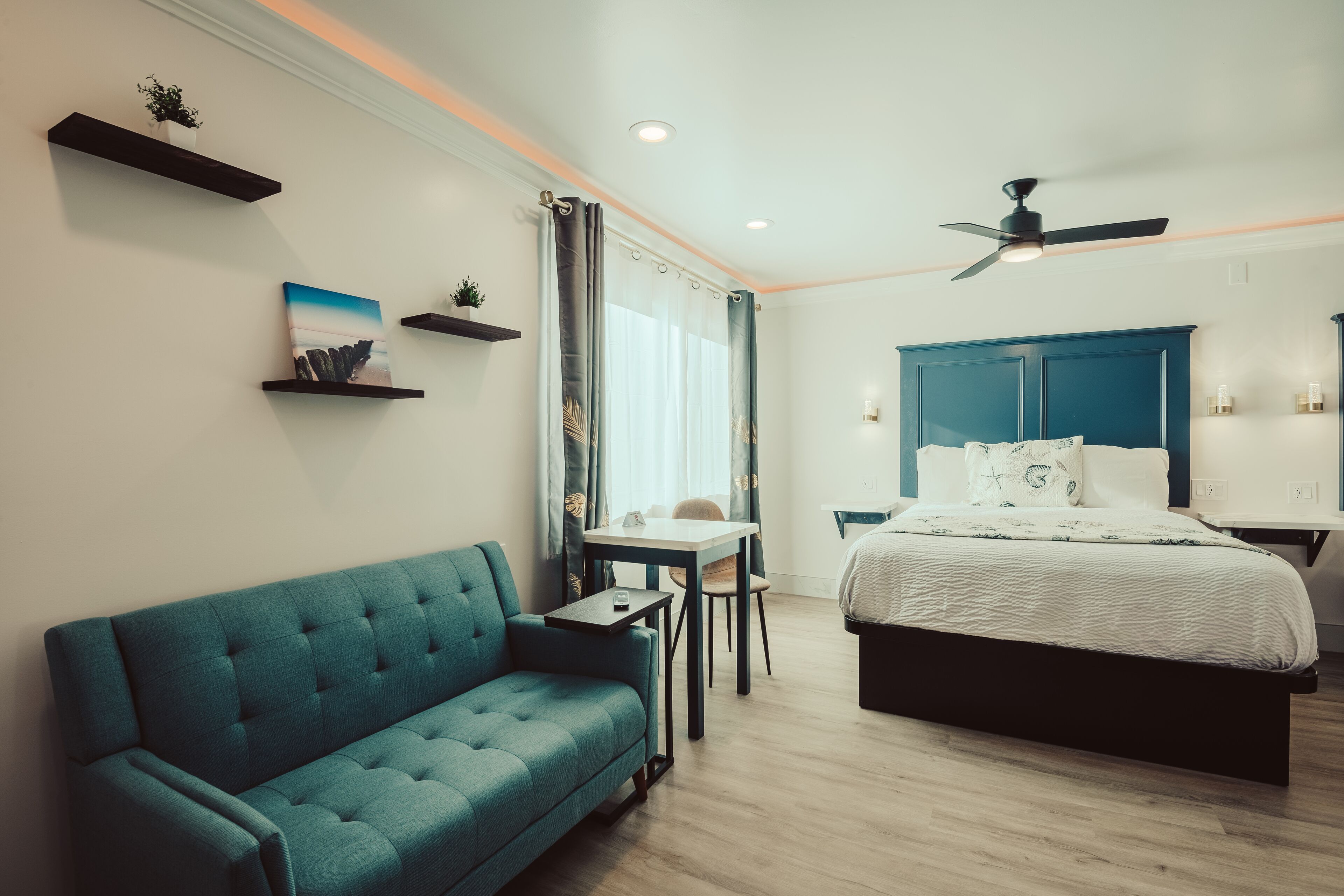 A modern bedroom with a teal sofa, a bed with a blue headboard, a small table, and a ceiling fan.